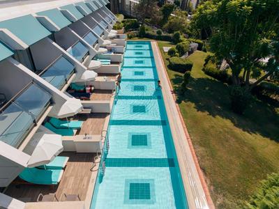 Vista di una lunga piscina all'aperto con lettini e ombrelloni accanto a un hotel e aree verdi.