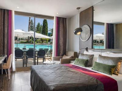 Chambre d'hôtel moderne avec grand lit, vue sur la piscine et le jardin à travers de grandes fenêtres.