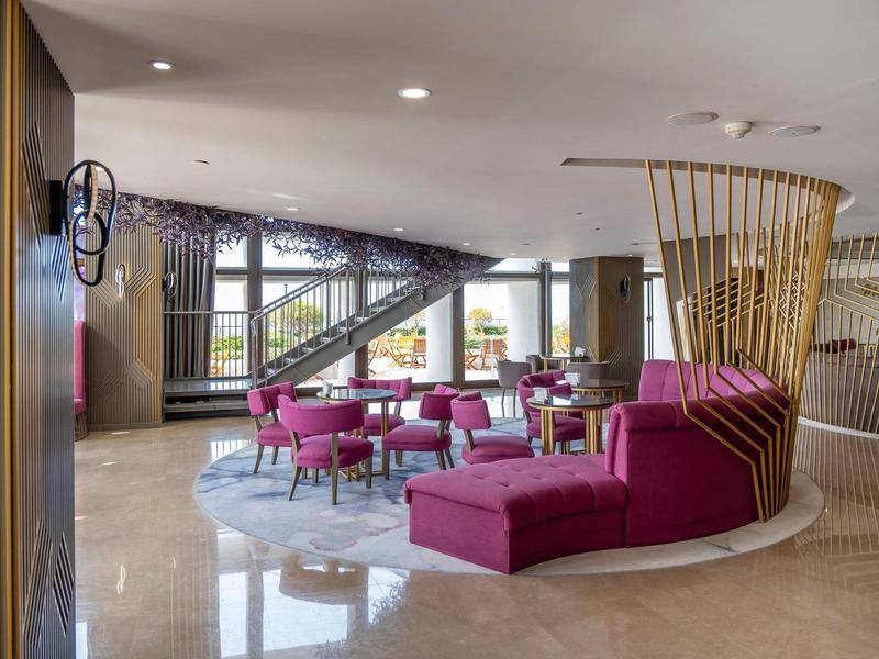 Area lounge moderna di hotel con sedie e divani rosa, pavimento chiaro e grandi finestre.