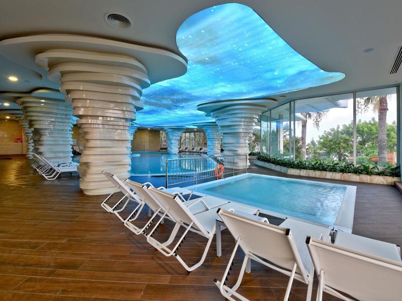 Area piscina interna moderna con lettini bianchi e colonne a forma organica e soffitto di vetro.