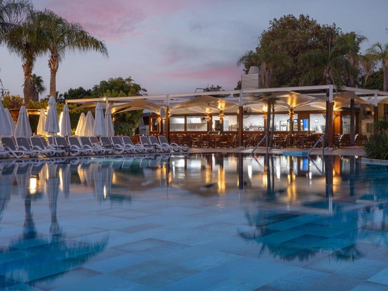 Hotel con ristorante illuminato e piscina al tramonto, circondato da palme e sedie a sdraio.