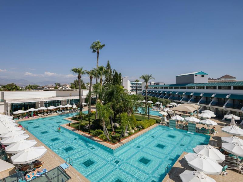 Piscina dell'hotel con palme e ombrelloni sotto un cielo sereno, circondata da edifici dell'hotel.