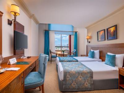 Camera d'hotel luminosa con due letti singoli, scrivania, sedia e balcone con vista mare.