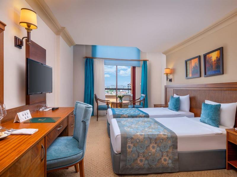 Camera d'hotel luminosa con due letti singoli, scrivania, sedia e balcone con vista mare.
