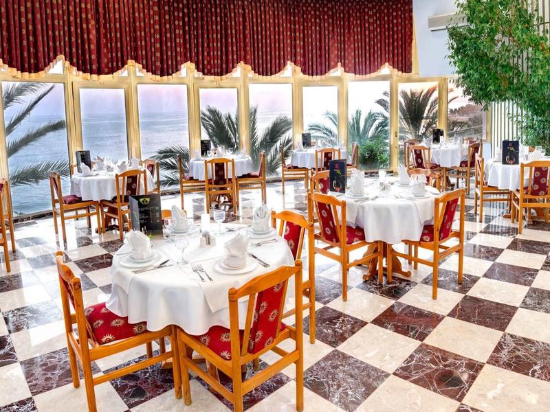 Restaurant élégant avec nappes, mobilier en bois et grandes fenêtres donnant sur des palmiers.