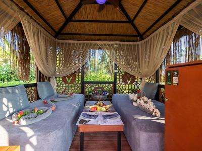 Sala acogedora con cojines azules, mesa y cortinas bajo un techo de madera en un entorno tropical.