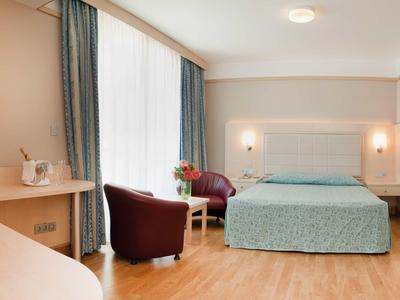 Helles Hotelzimmer mit Doppelbett, zwei Sesseln, Holzboden und großem Fenster mit blauen Vorhängen.