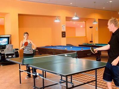Zwei Personen spielen Tischtennis in einem orangefarbenen Raum mit Tischen und Spielautomaten.