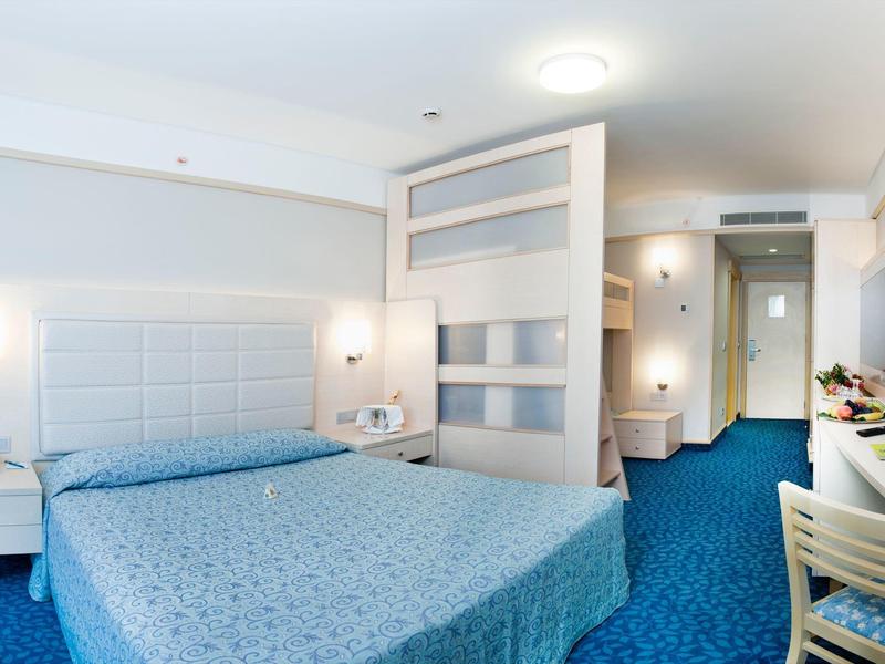Bright hotel room with large bed, blue carpet, and modern white furnishings.