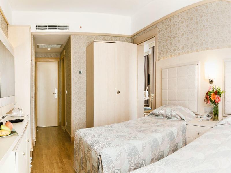 Bright hotel room with two beds, wooden floor, flower decoration, and TV on the wall.