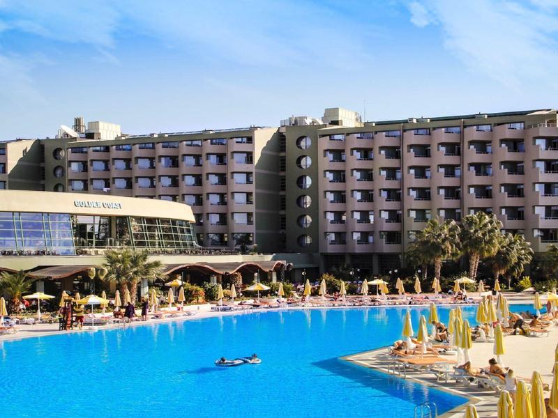 Large multi-story hotel with a big pool in the foreground under a blue sky.
