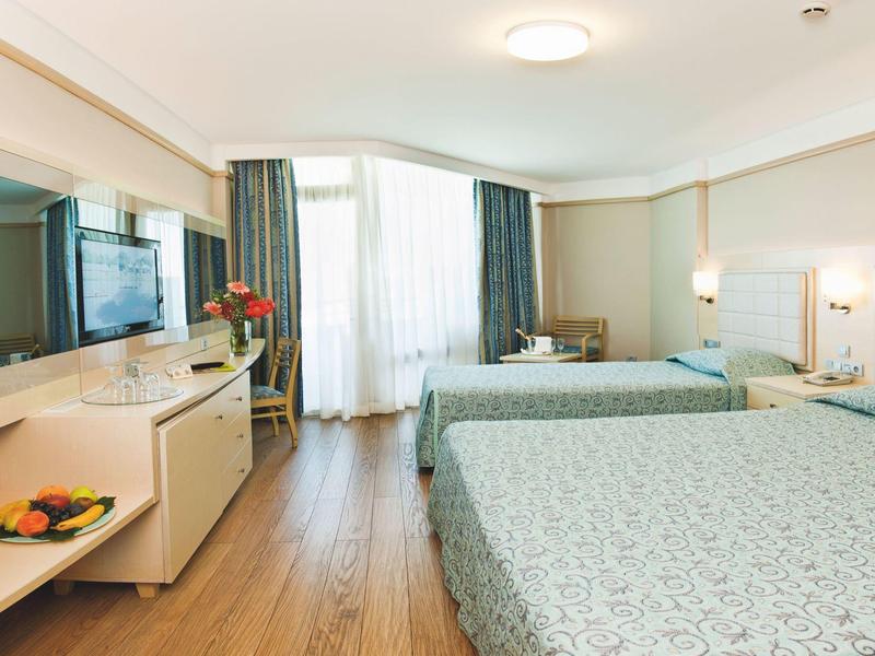 Bright hotel room with two beds, TV, desk, and large windows with curtains.