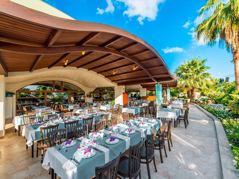 Outdoor restaurant with set tables under a curved wooden roof next to palm trees.
