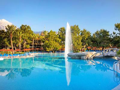 Groot blauw hotelzwembad met fontein, omgeven door bomen en ligstoelen op een zonnige dag.