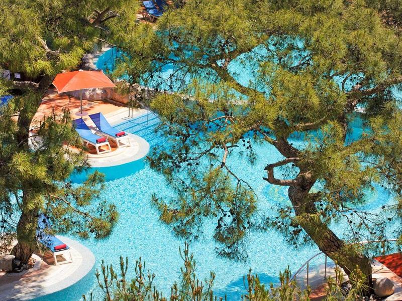 Zwembad met blauwe ligbedden, parasols en bomen bij een hotel of resort.