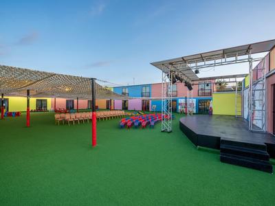 Area esterna colorata con prato, palco e aree coperte per sedersi in un villaggio turistico