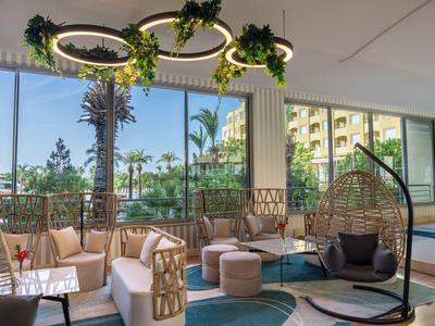 Heller Hotel-Loungebereich mit Sitzmöbeln, Hängekorb und großen Fenstern mit Blick auf Palmen.