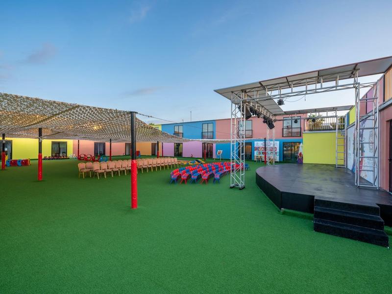 Area esterna colorata con prato, palco e aree coperte per sedersi in un villaggio turistico