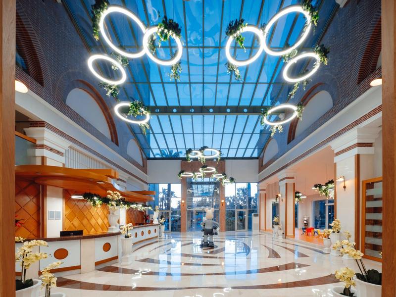 Ampia hall di hotel luminosa con tetto in vetro e lampade ad anello moderne sul soffitto.