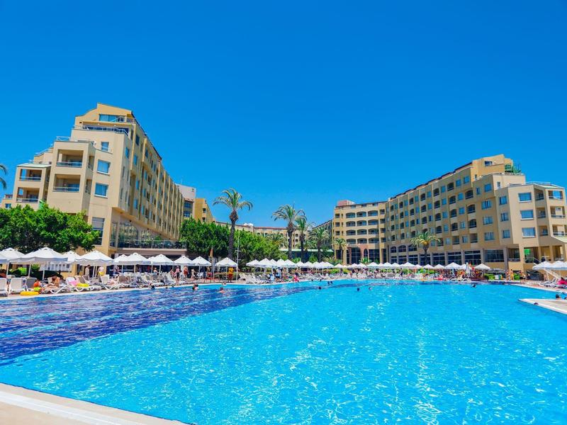 Grande piscina all'aperto davanti a due edifici alberghieri a più piani sotto un cielo azzurro limpido.