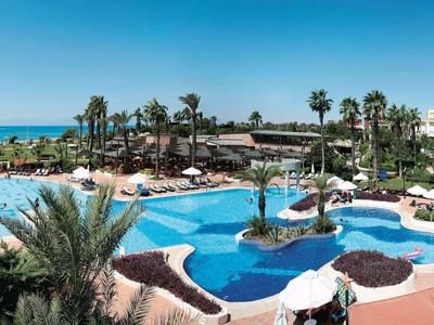 Panoramaansicht eines Resorts mit Pool, Palmen, Sonnenschirmen und blauem Himmel am Meer.