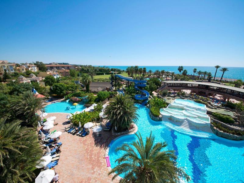 Complesso alberghiero con piscine, palme e vista sul mare sotto un cielo azzurro.