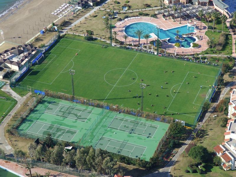 Veduta aerea di un resort con campo da calcio, campi da tennis, piscina e spiaggia vicina.