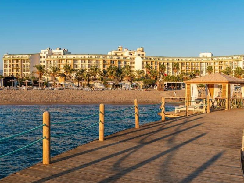 Vista di un grande hotel sulla spiaggia con una piccola passerella in primo piano sotto un cielo sereno.