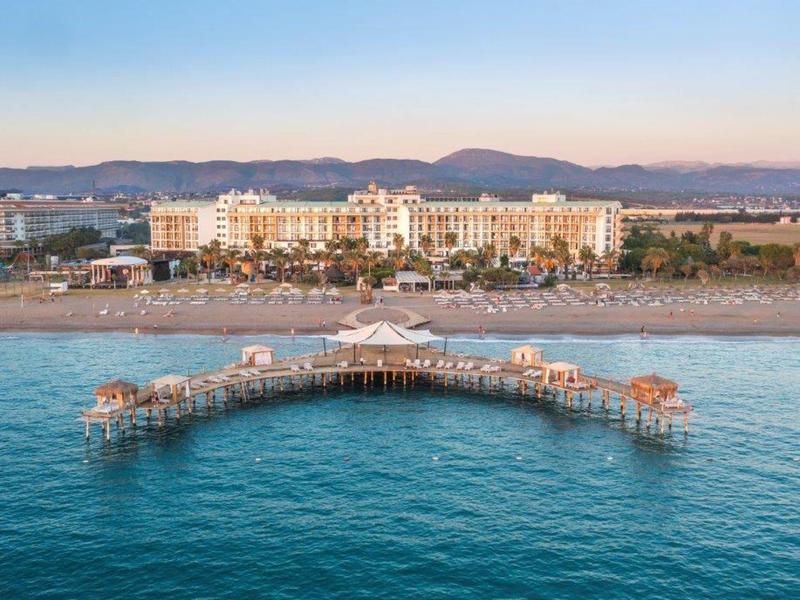 Hotel con spiaggia e molo che si estende nel mare blu calmo, montagne sullo sfondo sotto un cielo sereno.