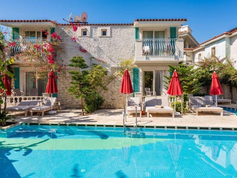 Hotel con facciata in pietra, balconi, piscina e ombrelloni rossi sotto un cielo blu.