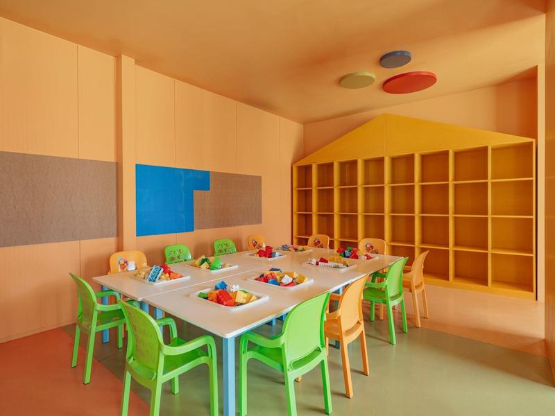 Stanza colorata dell'asilo con tavoli, sedie e scaffali per giocattoli e materiali.