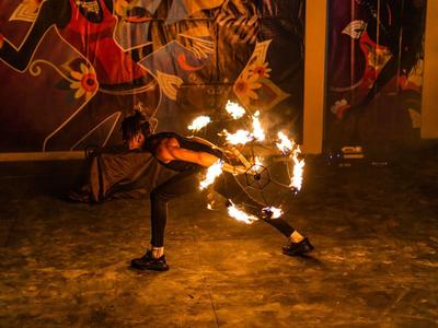 Feuerkünstler in dunkler Kleidung führt Flammenjonglage in einem schummrigen Raum mit Wandkunst auf.