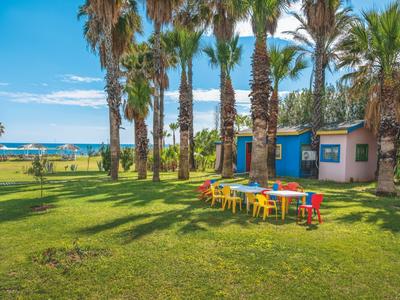 Grüner Garten mit Palmen, bunten Tischen und Stühlen, blauer Himmel und Meer im Hintergrund.