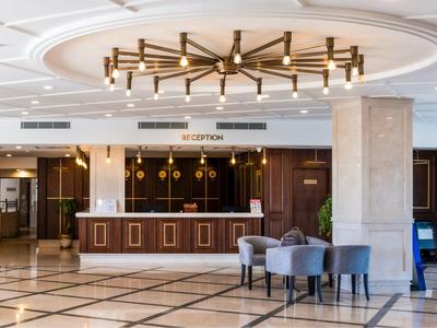 Heller Hotellobby mit modernem Kronleuchter, Empfangstresen aus Holz und grauen Sesseln.
