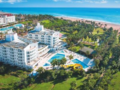 Großes weißes Hotel mit Pool, umgeben von grünen Bäumen und Blick auf den Strand und das Meer.