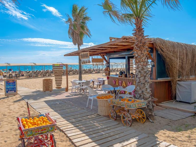 Chiosco sulla spiaggia con palme, tavoli e sedie su sabbia sotto cielo blu.