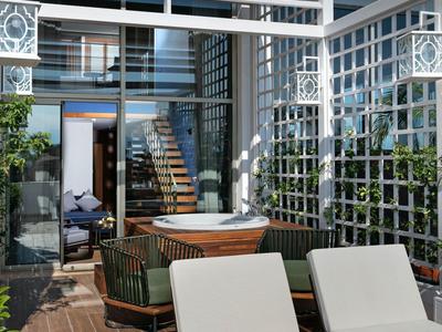 Area lounge esterna moderna con sedie imbottite, tavolo in legno e scala sotto un tetto di vetro.