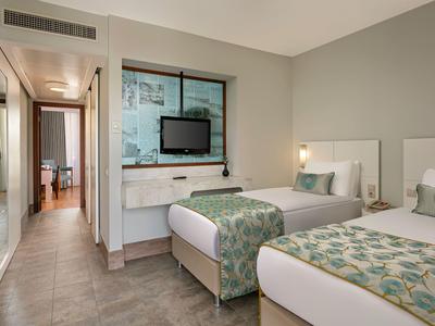 Camera d'hotel moderna con due letti singoli, TV a muro e corridoio verso l'ingresso.