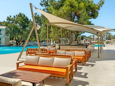 Area salotto soleggiata a bordo piscina con vele ombreggianti, cuscini e tavoli in legno vicino a piscina limpida.