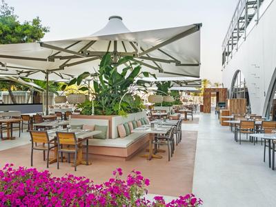 Area pranzo esterna dell'hotel con tavoli, sedie, ombrelloni e piante in una giornata di sole.