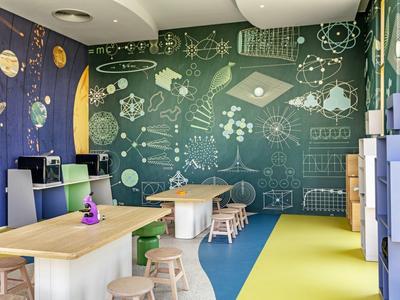 Stanza luminosa per bambini con decorazioni murali a tema spazio, tavoli, sgabelli e armadietti.