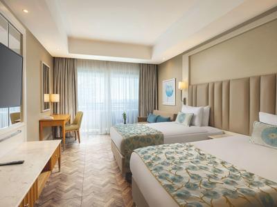 Camera d'hotel moderna con due letti, scrivania, TV e ampia finestra con tende.