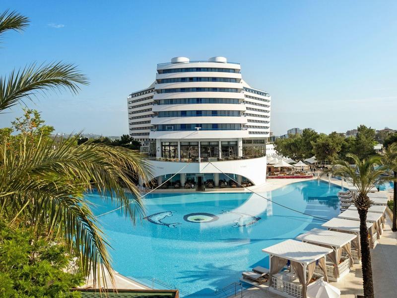 Hotel moderno con grande piscina circolare circondata da palme e lettini sotto un cielo azzurro.