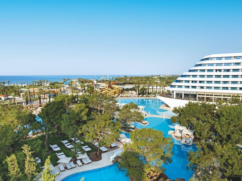 Resort con piscina, alberi, lettini e un moderno edificio dell'hotel bianco sotto un cielo azzurro.