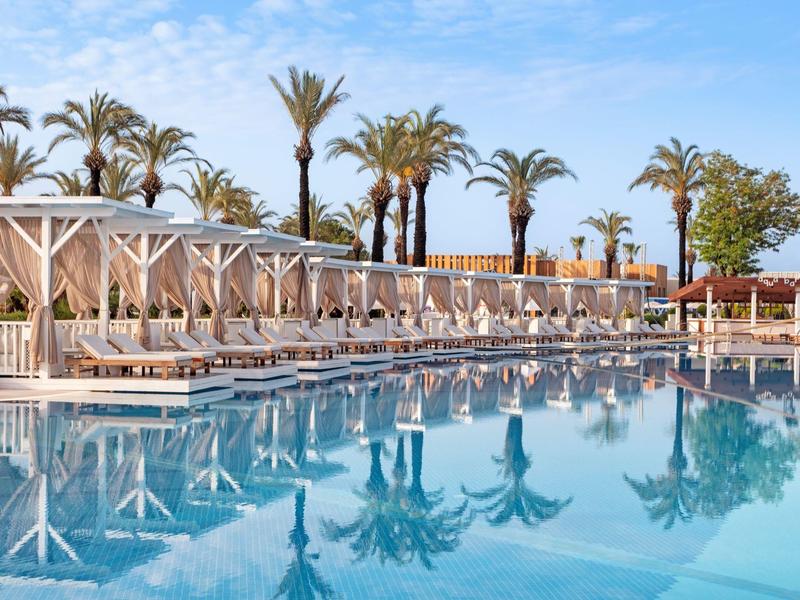 Piscina all'aperto con lettini, ombrelloni e palme sotto un cielo azzurro.