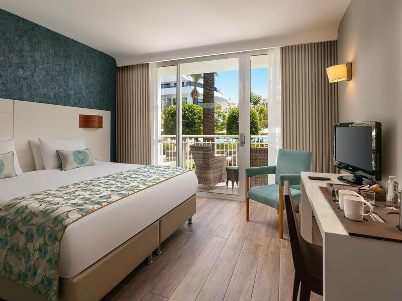 Camera d'hotel luminosa con letto matrimoniale, balcone, scrivania, TV e area salotto.