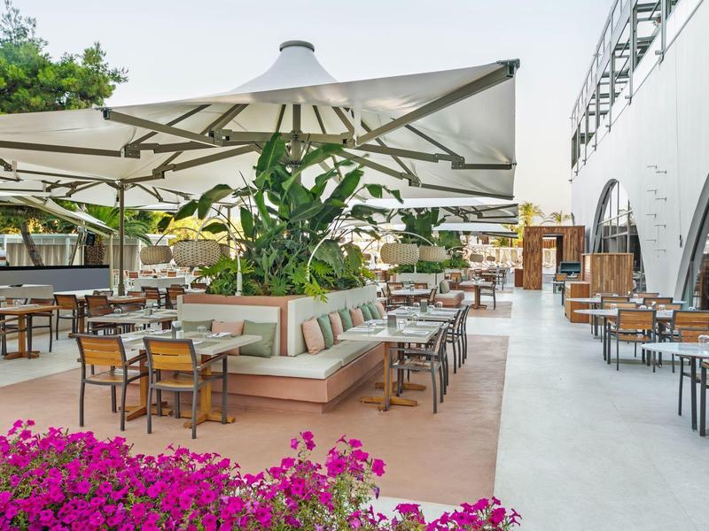 Area pranzo esterna dell'hotel con tavoli, sedie, ombrelloni e piante in una giornata di sole.