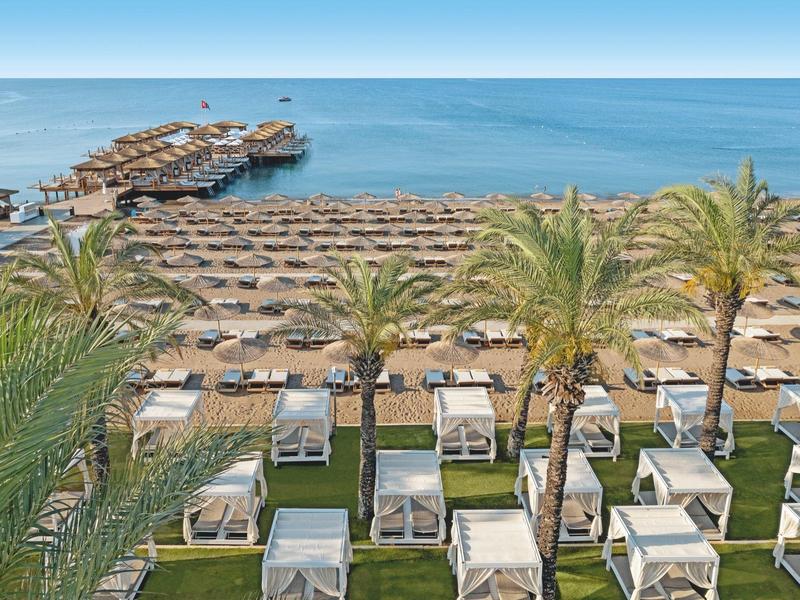 Resort sulla spiaggia con cabine, lettini, palme e molo di legno che si estende sul mare.