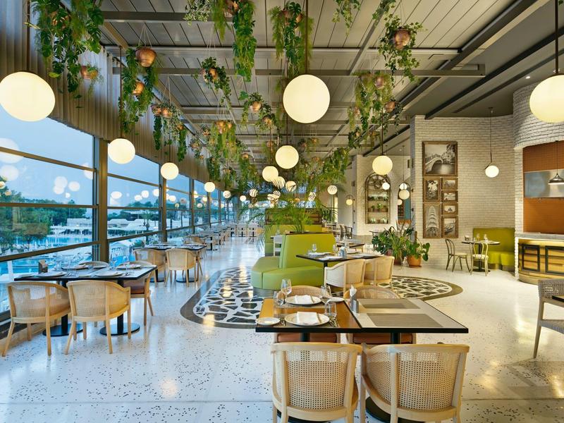 Ristorante moderno con piante sospese, luci rotonde e grandi finestre che si affacciano sull'acqua.