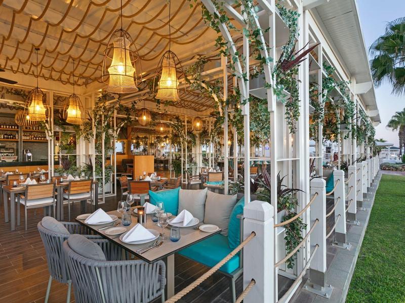 Zona pranzo esterna con sedie in vimini, cuscini turchesi e lanterne sospese sotto un pergolato.